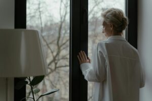 A senior woman in white looking out a window thoughtfully, symbolizing reflection.