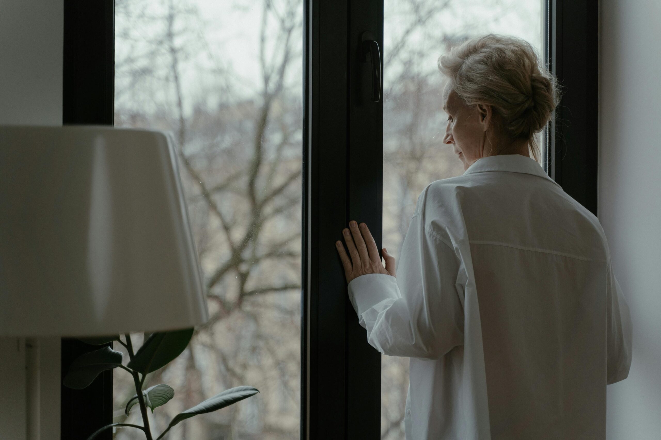 A senior woman in white looking out a window thoughtfully, symbolizing reflection.