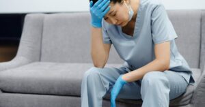 Tired nurse in scrubs sitting on a sofa, looking exhausted with her hand on her head.